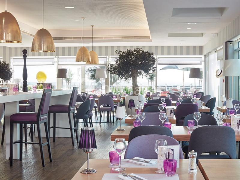 Salle de restaurant lumineuse avec tables dressées, chaises violettes, luminaires dorés et grandes baies vitrées.