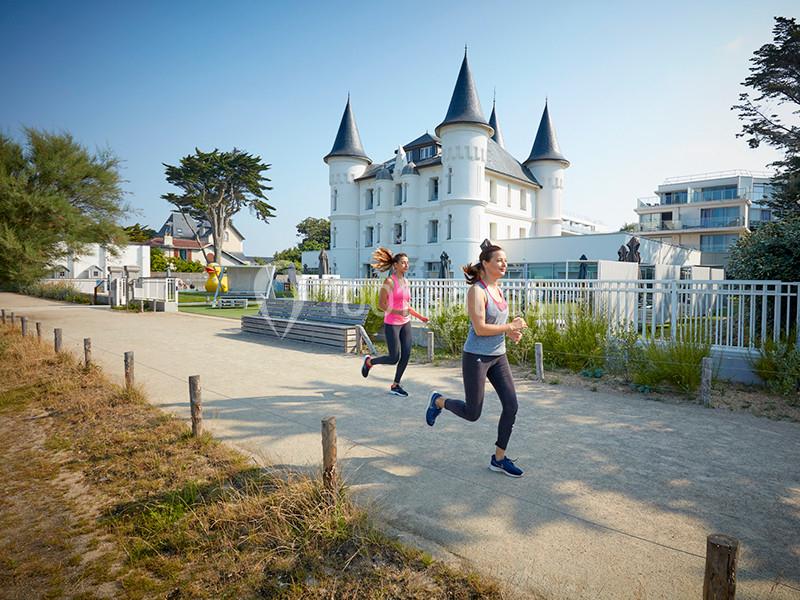 Deux femmes courent sur un chemin bordé de verdure, avec un château blanc en arrière-plan par temps ensoleillé.
