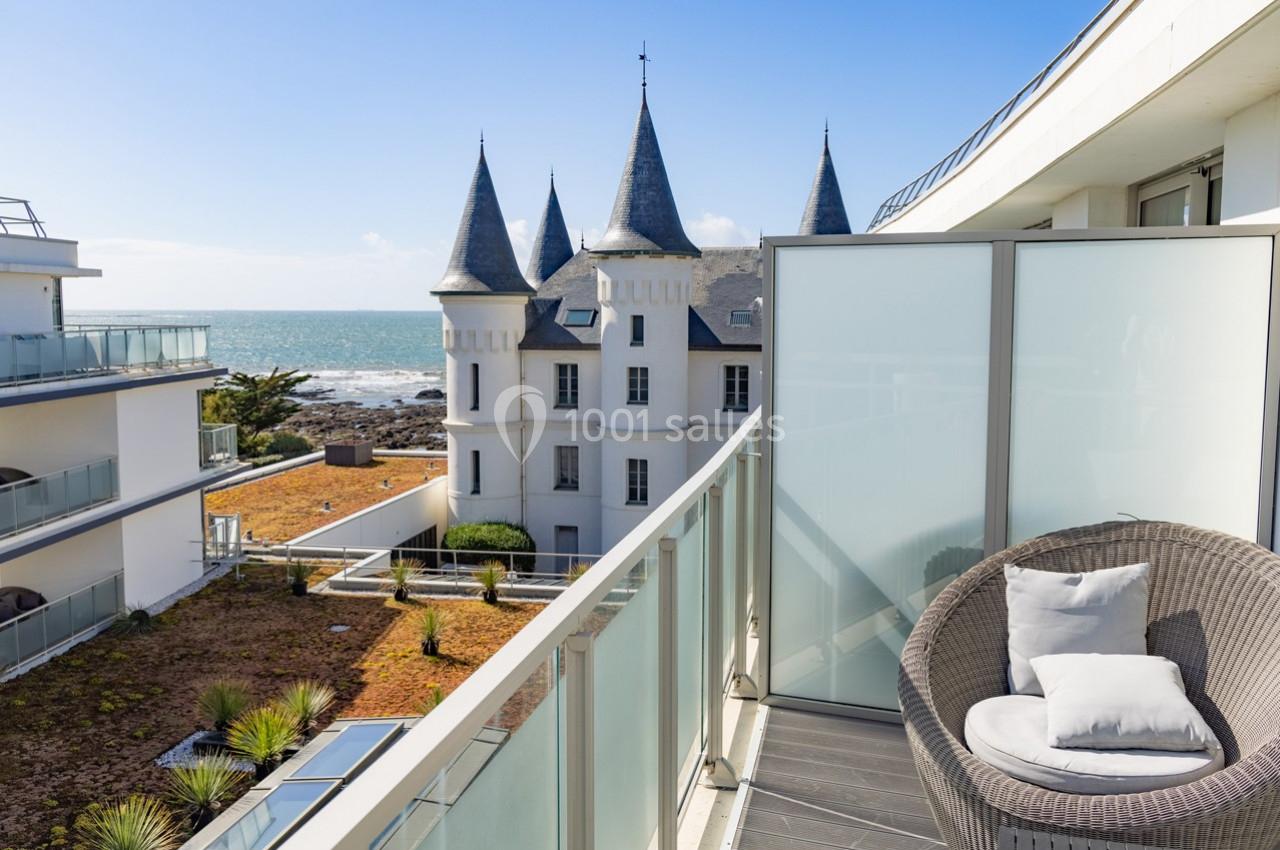 Vue depuis un balcon moderne avec fauteuil, donnant sur un château en bord de mer et des bâtiments contemporains.