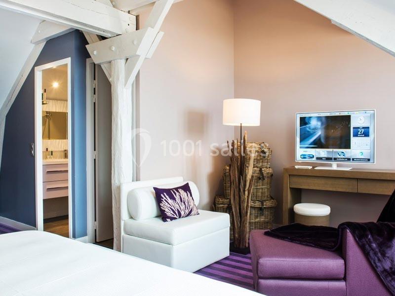 Chambre lumineuse avec poutres apparentes, fauteuils modernes, bureau avec écran et vue sur une salle de bain ouverte.