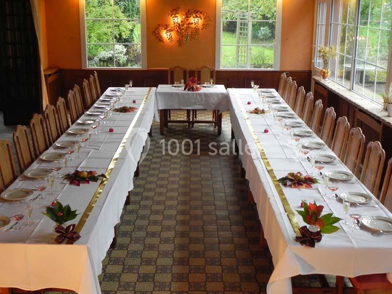 Salle de réception avec deux longues tables dressées, ornées de décorations florales, près de grandes fenêtres donnant sur…