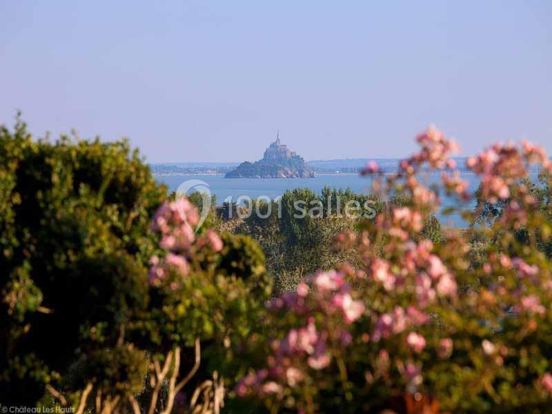 Vue du Mont-Saint-Michel au loin, entouré d'eau, avec des arbres et des fleurs roses au premier plan.