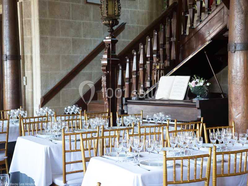 Salle élégante avec tables dressées, chaises dorées, piano ouvert et escalier en bois dans un cadre raffiné.