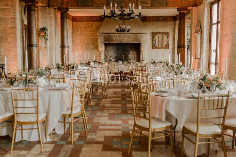 Salle de réception élégante avec tables rondes décorées de fleurs et chaises dorées, éclairée par des fenêtres et un lustre.