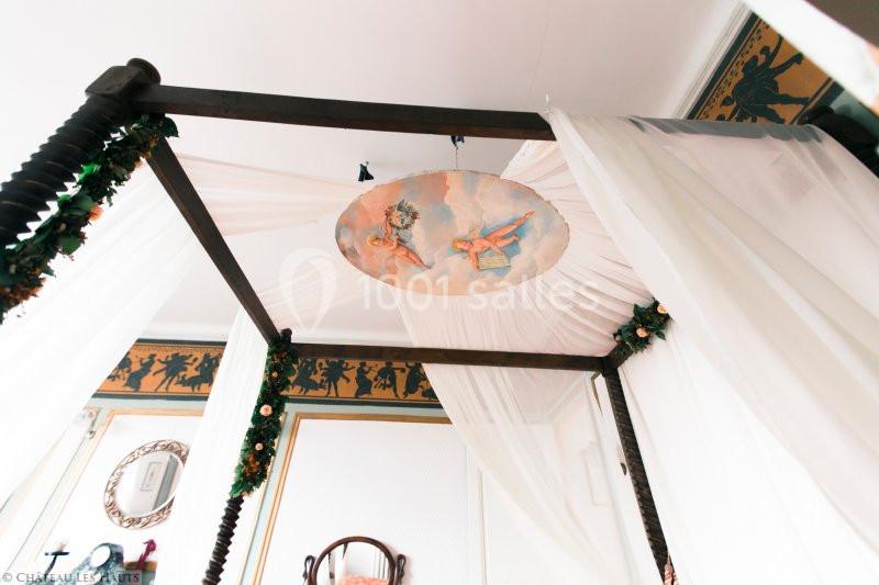 Lit à baldaquin en bois sombre décoré de voilages blancs et de guirlandes, avec un plafond peint au motif floral.