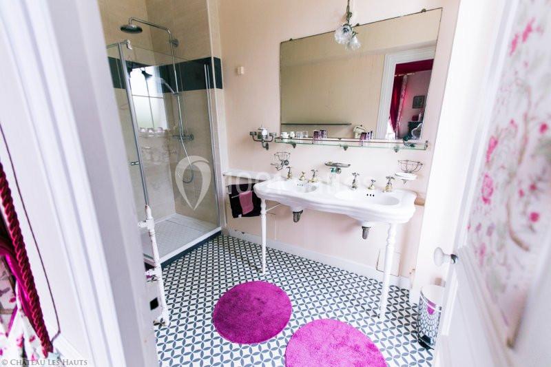 Salle de bain lumineuse avec douche vitrée, double vasque blanche, carrelage géométrique et tapis ronds roses.