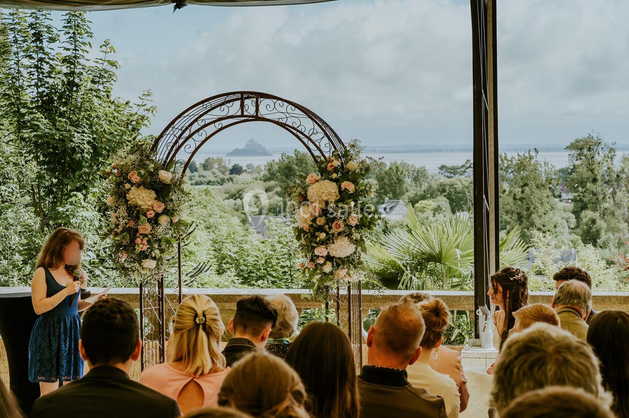 Une cérémonie en extérieur avec une arche fleurie, vue sur un paysage verdoyant et le Mont-Saint-Michel au loin.