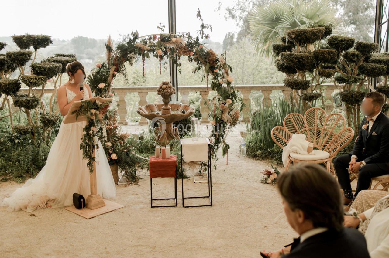 Une femme en robe de mariée lit un texte devant un autel décoré de fleurs, entourée d'invités assis.