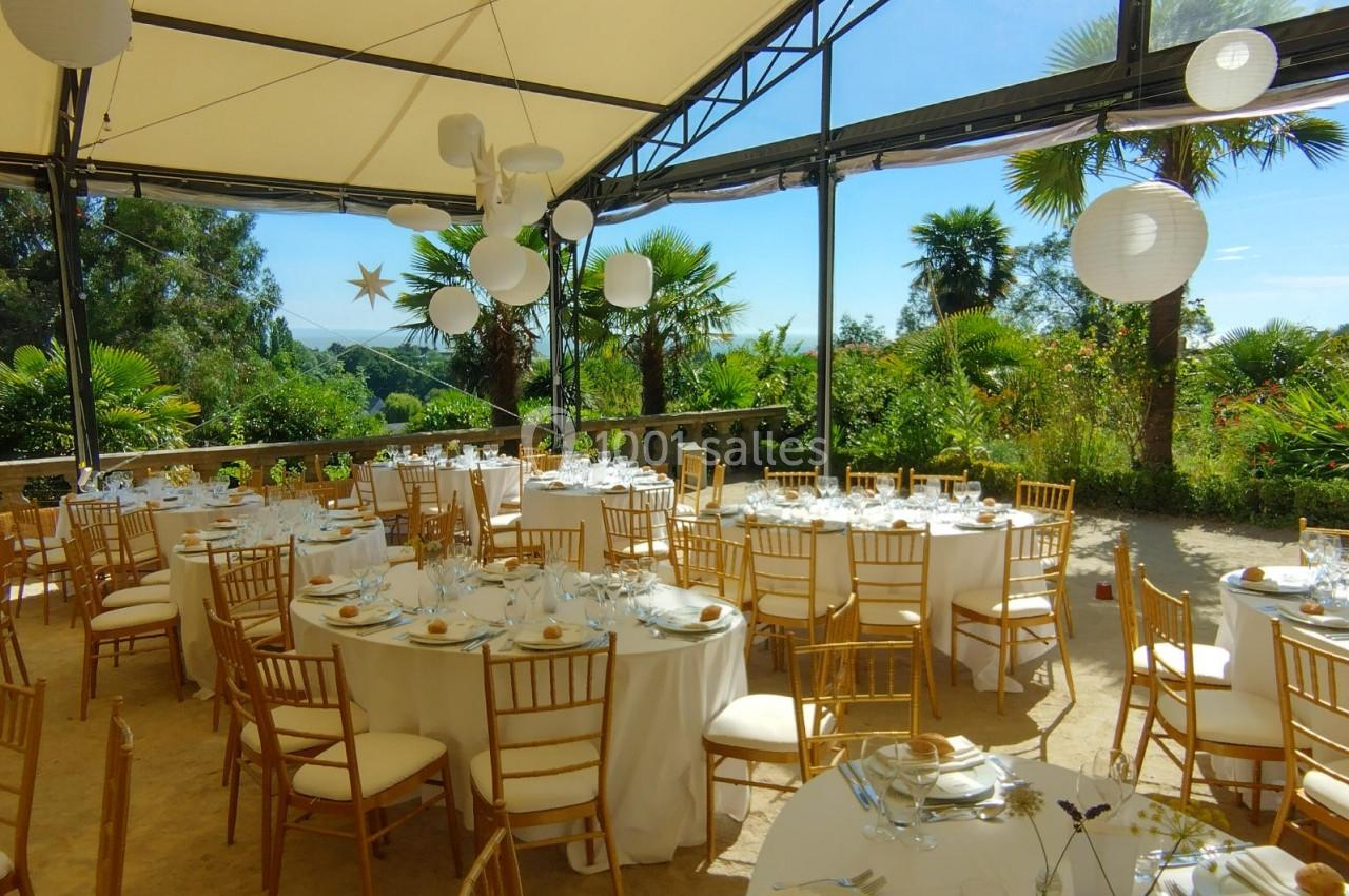 Salle de réception en plein air avec tables rondes dressées, chaises dorées et vue sur un jardin tropical.