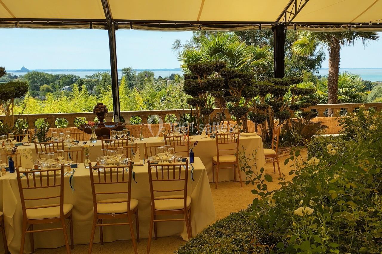 Salle de réception en plein air avec tables dressées, chaises dorées et vue sur un jardin arboré et la mer au loin.