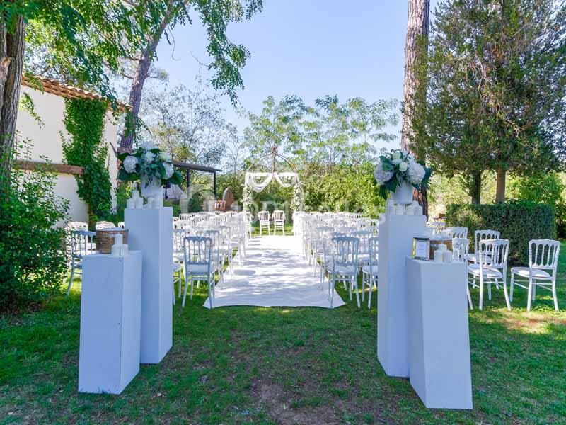 Allée centrale décorée pour une cérémonie de mariage en extérieur, entourée de chaises blanches et de verdure.