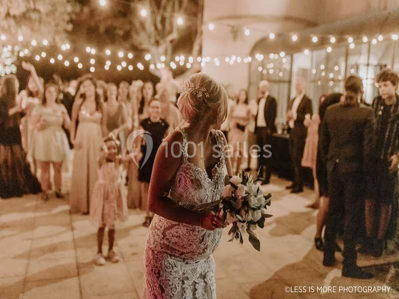 Une mariée en robe blanche tient un bouquet, entourée d'invités lors d'une réception en soirée éclairée par des guirlandes.