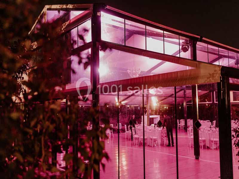 Salle de réception vitrée éclairée en rose, avec des tables dressées et quelques personnes debout à l'intérieur.