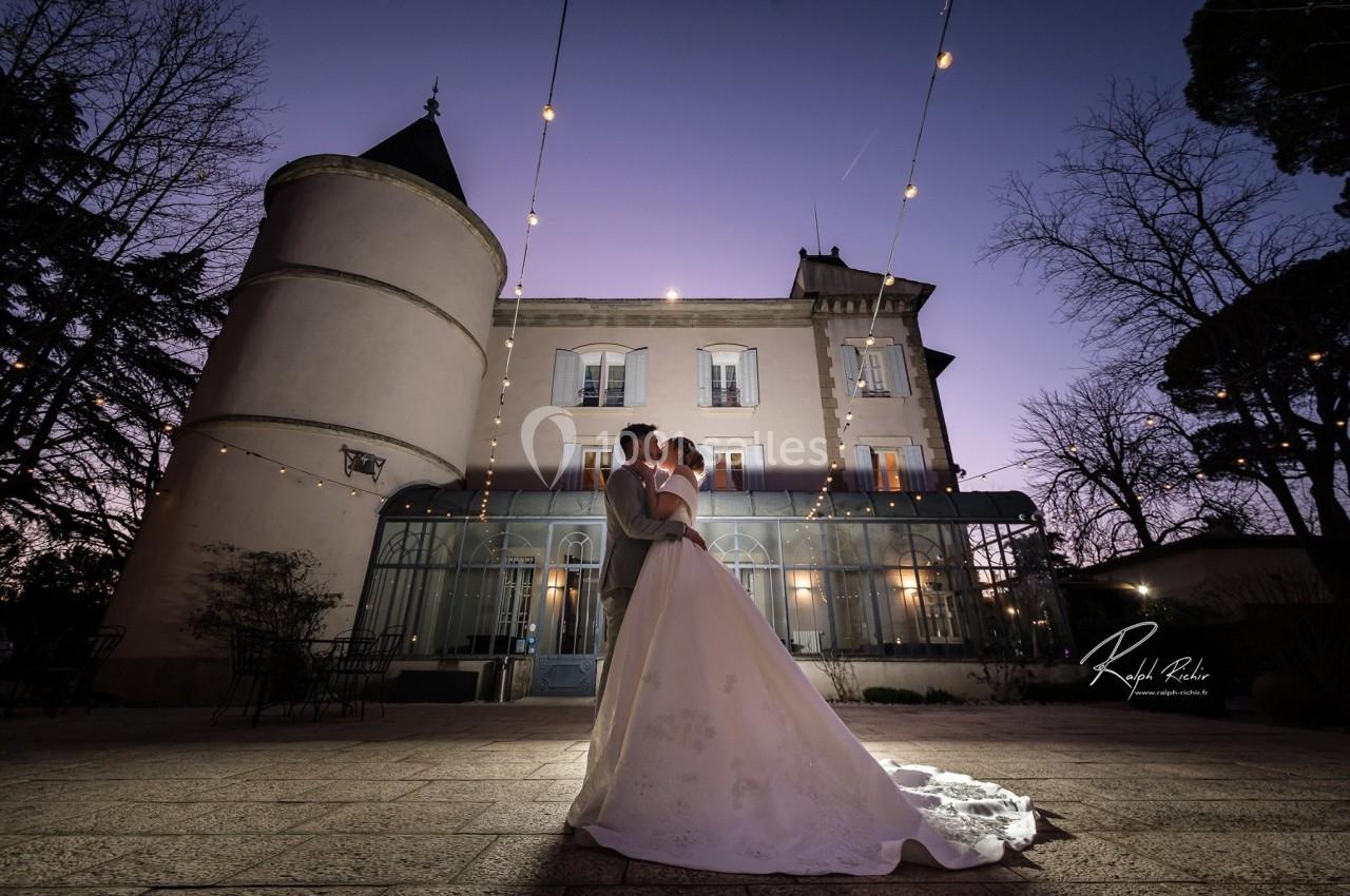 Un couple en tenue de mariage pose devant un château éclairé au crépuscule, entouré de guirlandes lumineuses.