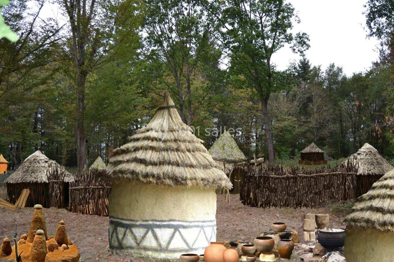 Village traditionnel avec huttes en toit de chaume entourées de palissades en bois, au milieu d'une forêt dense.
