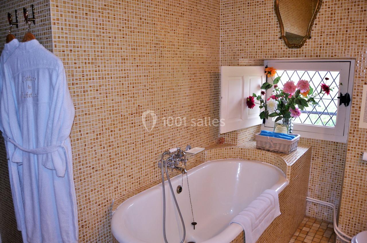 Salle de bain avec baignoire blanche, murs en mosaïque beige, fenêtre avec grille et vase de fleurs colorées.
