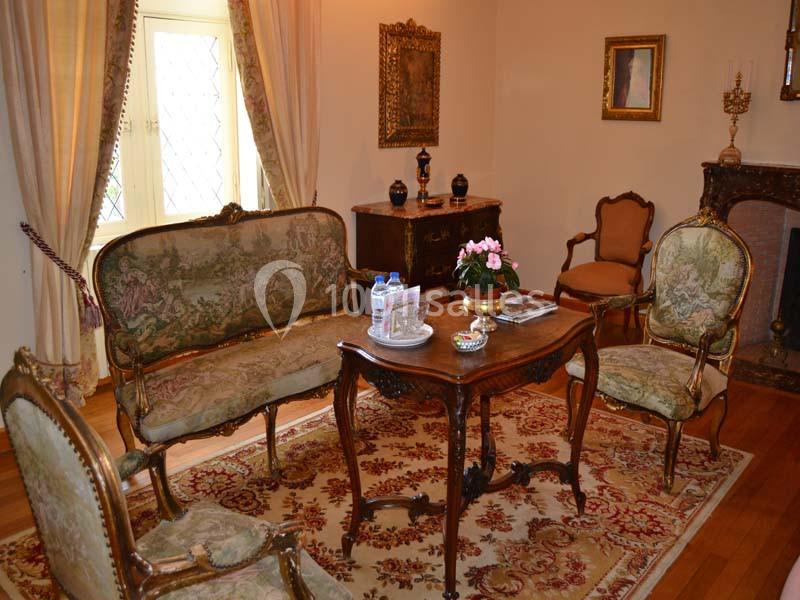 Salon ancien avec mobilier en bois sculpté, tapis floral, table centrale et décoration classique sur fond de murs clairs.