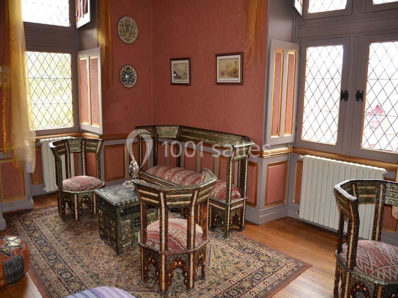 Salon décoré de meubles en bois sculpté et incrusté, tapis oriental au sol, murs rouges ornés de cadres et assiettes.