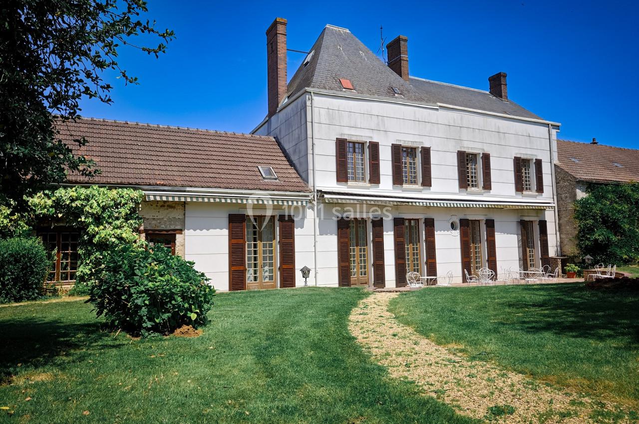 Grande maison blanche à deux étages avec volets marron, entourée d'un jardin verdoyant et d'un chemin en gravier.
