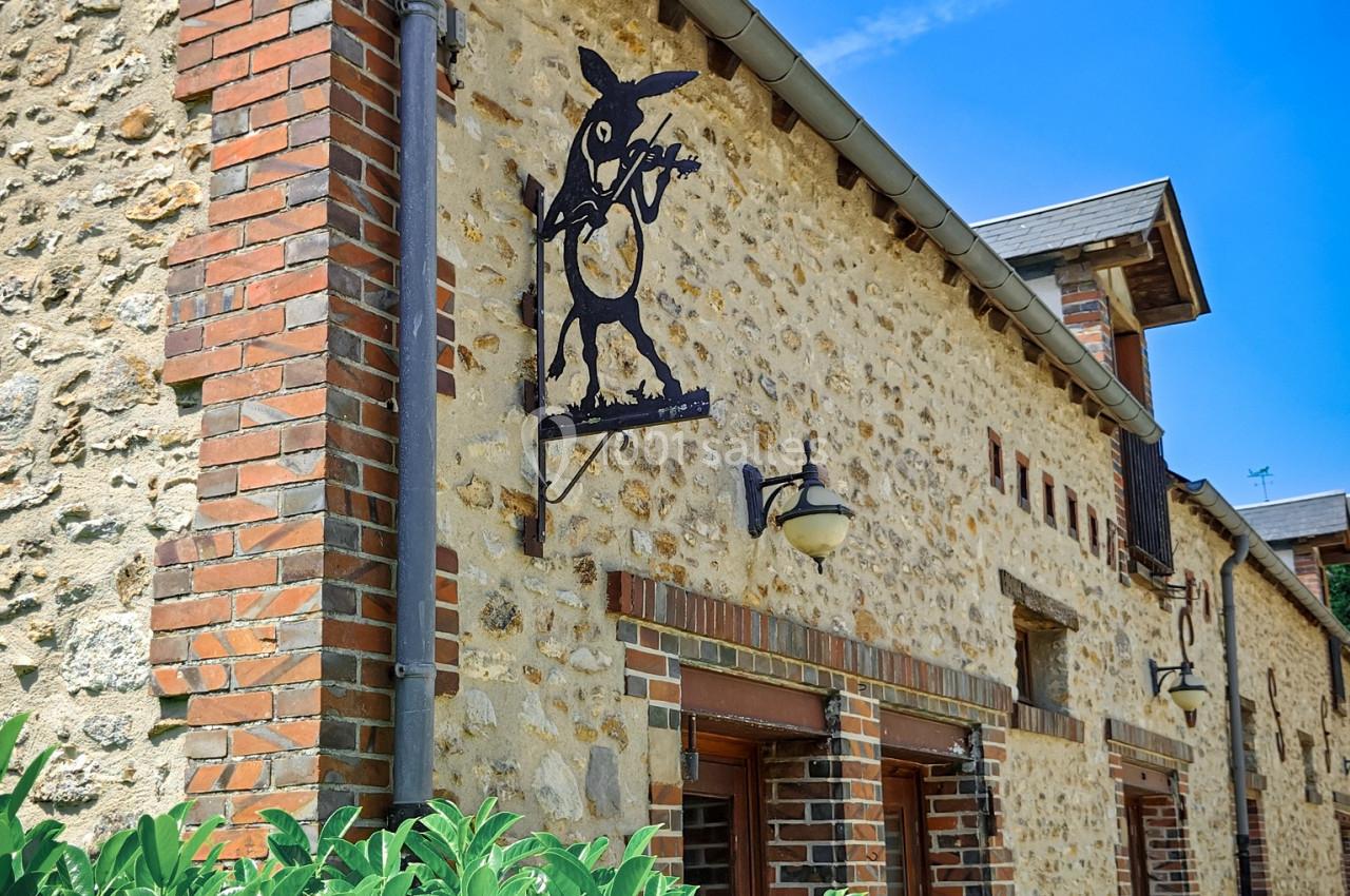Façade en pierre d'une maison avec enseigne métallique décorative et buisson au premier plan, sous un ciel bleu.
