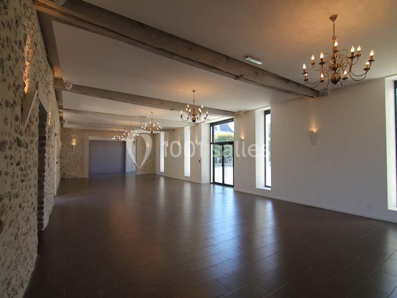 Salle spacieuse avec murs en pierre, poutres apparentes, lustres suspendus et grandes fenêtres laissant entrer la lumière.