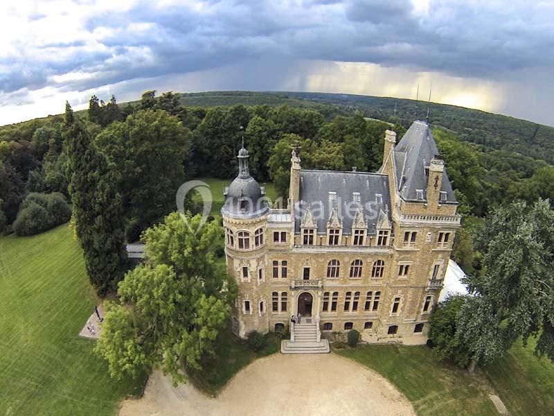 Vue aérienne d'un château en pierre entouré de pelouses, d'arbres et d'une forêt sous un ciel nuageux.