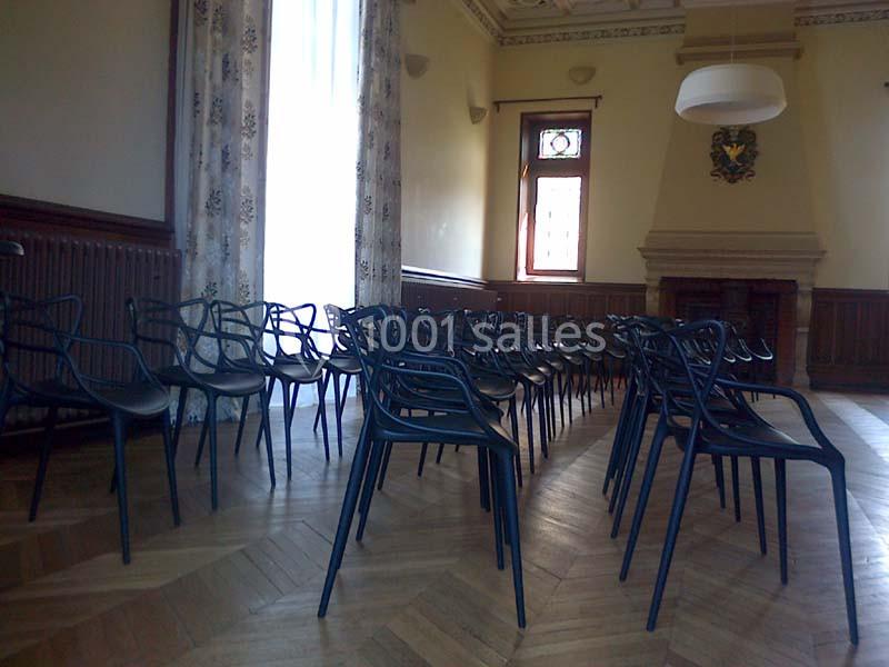 Salle lumineuse avec des rangées de chaises noires alignées sur un parquet, près de grandes fenêtres et d'une cheminée.