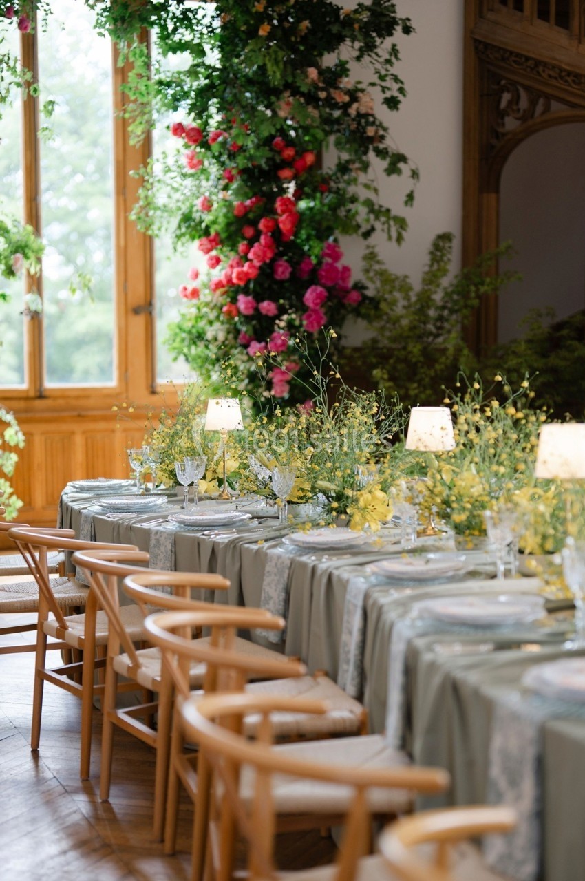 Tables élégamment dressées avec nappes vertes, chaises en bois et décor floral abondant dans une salle lumineuse.