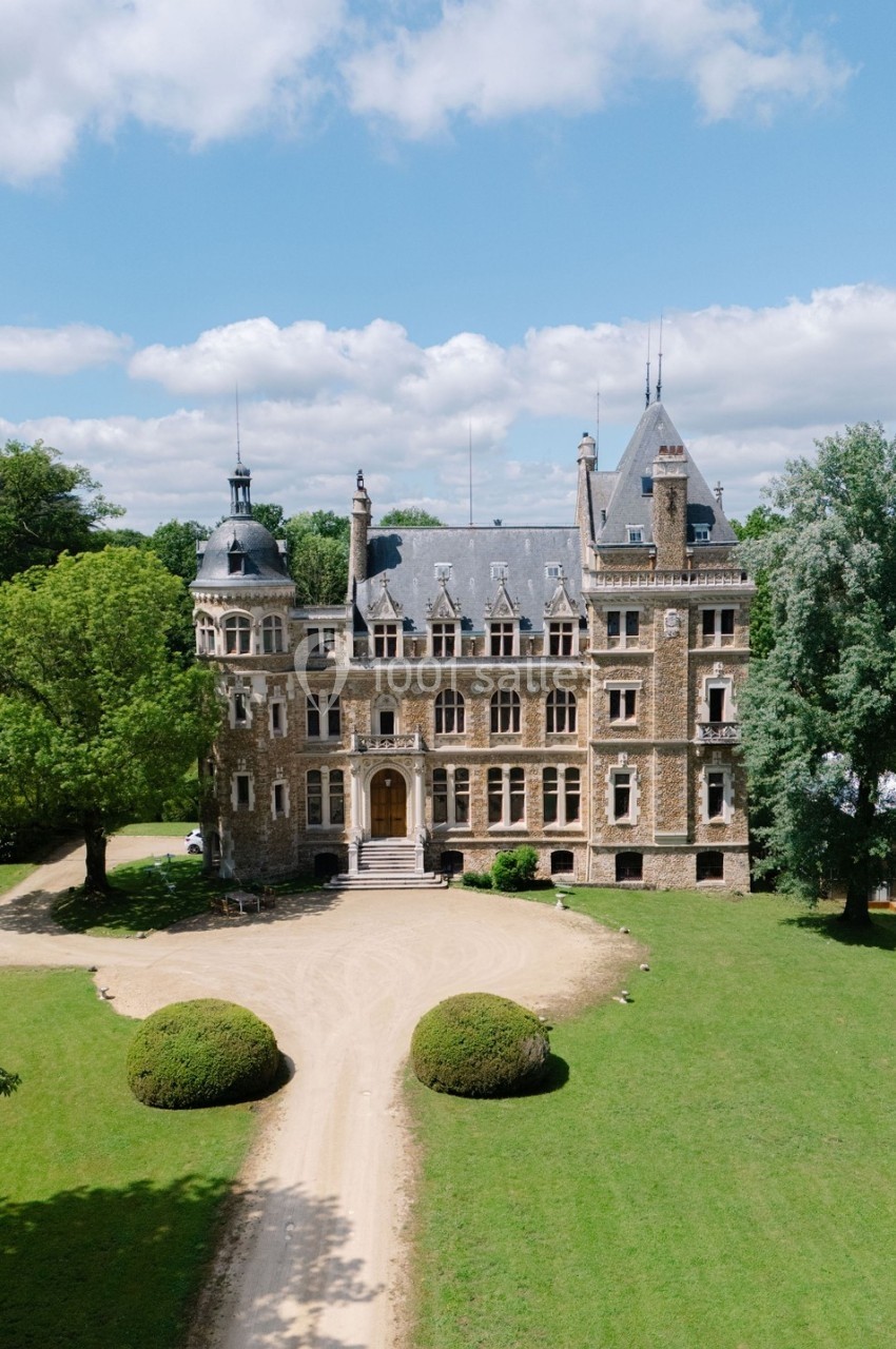 Château en pierre entouré de verdure, vu depuis une allée centrale sous un ciel partiellement nuageux.