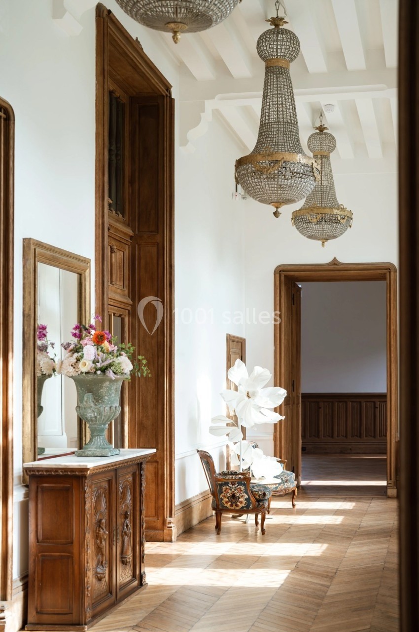 Un couloir lumineux avec parquet, meubles en bois sculpté, lustres en cristal et décoration florale.