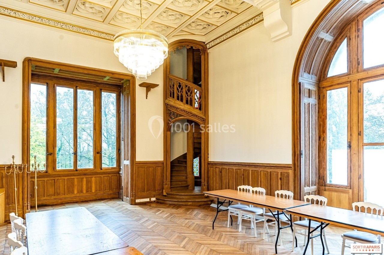 Salle lumineuse avec parquet, boiseries, grande fenêtre, lustre et escalier en colimaçon sculpté en bois.