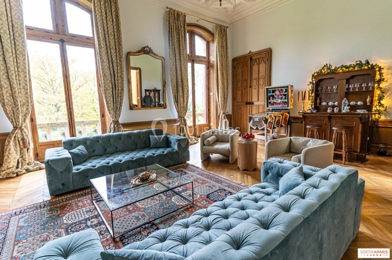 Salon élégant avec canapés en velours bleu, table basse en verre, tapis coloré et meubles anciens près de grandes fenêtres.