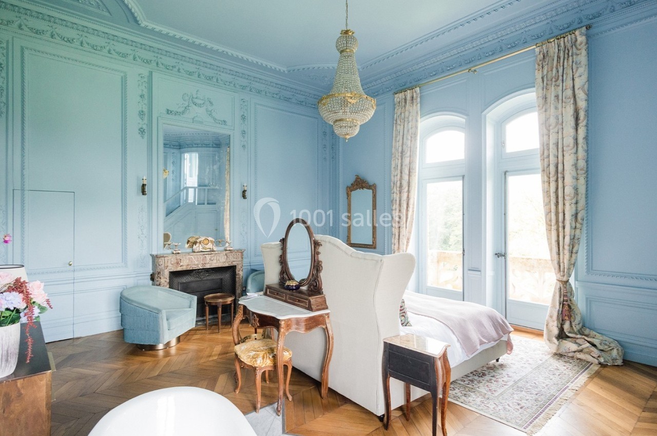 Chambre élégante avec murs bleus, lit double, cheminée, lustre et grandes fenêtres donnant sur un jardin.