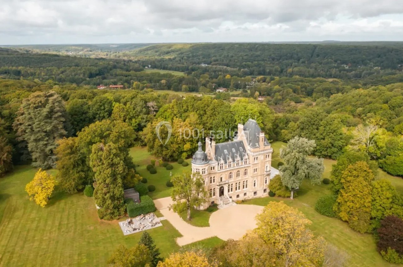 Vue aérienne d'un château entouré de vastes jardins et de forêts sous un ciel partiellement nuageux.