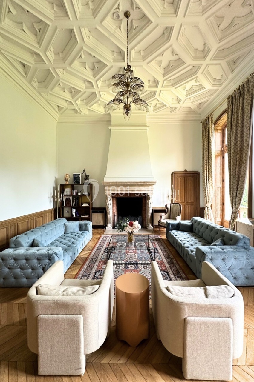 Salon spacieux avec plafond orné, cheminée centrale, canapés bleus, fauteuils beiges et tapis persan au sol.