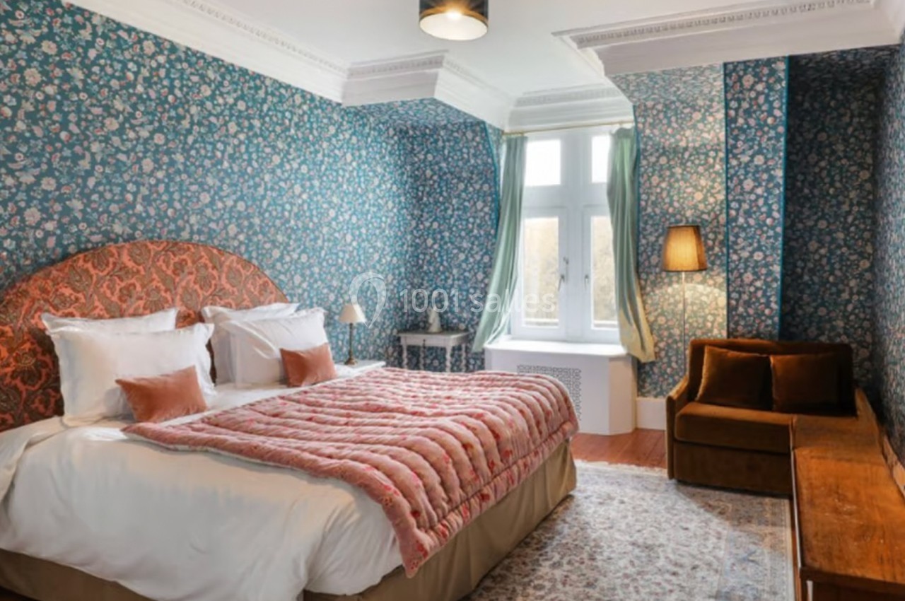 Chambre avec lit double, tête de lit rouge, murs à motifs floraux, canapé marron et grande fenêtre avec rideaux verts.