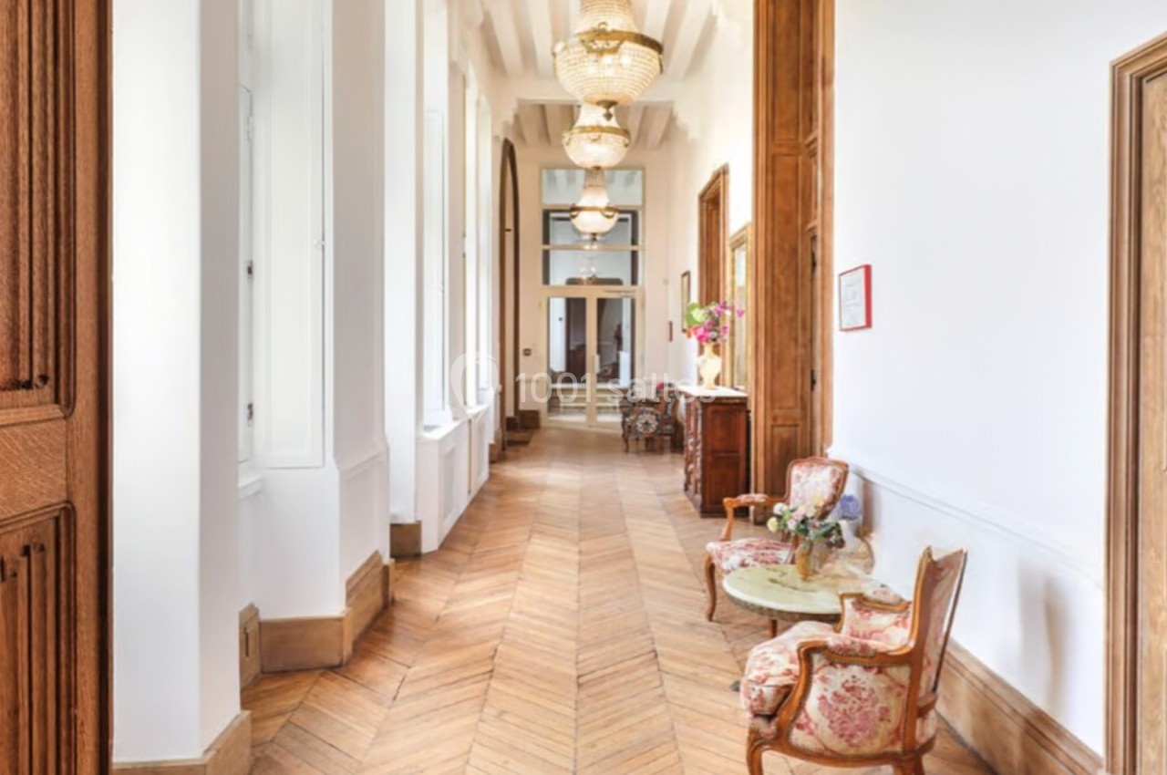 Un couloir lumineux avec parquet en bois, murs blancs, lustres suspendus et mobilier ancien près des fenêtres.
