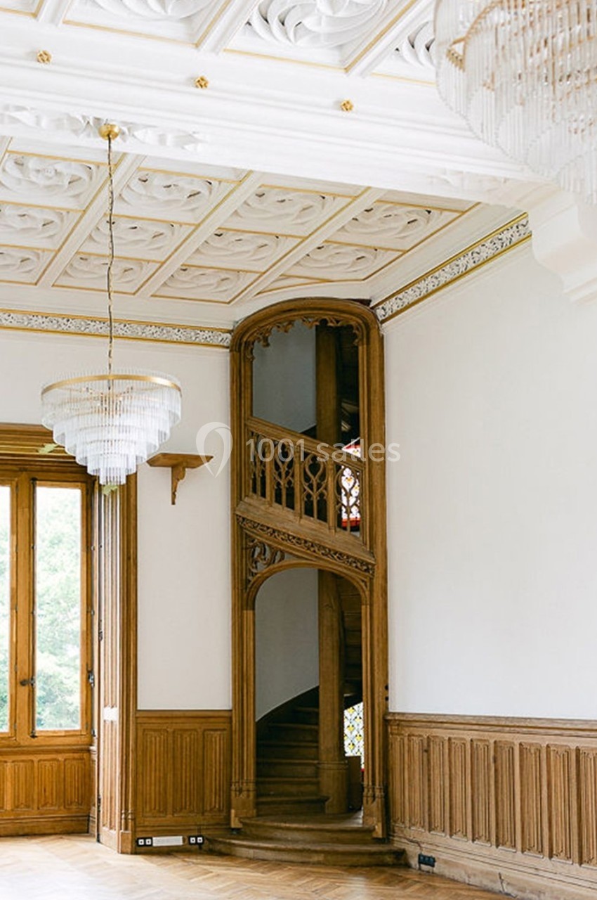 Escalier en bois sculpté dans une pièce lumineuse avec plafond orné et lustres en cristal.