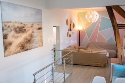 Salle de réception avec murs en briques, poutres apparentes, tables hautes et chaises pliantes sous des lampes suspendues.