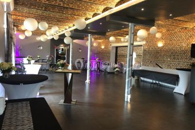 Salle de réception avec murs en briques, poutres apparentes, tables hautes et chaises pliantes sous des lampes suspendues.