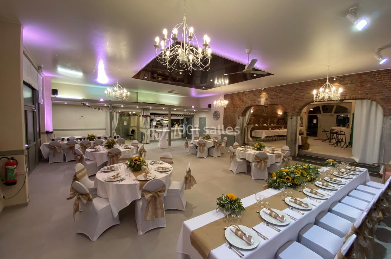 Salle de réception décorée avec des tables rondes et rectangulaires, nappes blanches, chaises ornées et éclairage chaleureux.