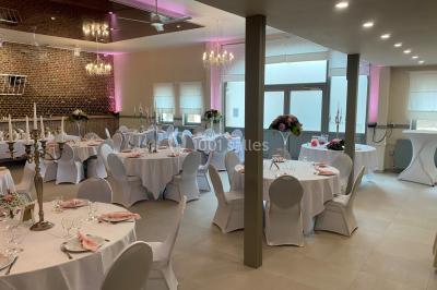 Salle de réception avec murs en briques, poutres apparentes, tables hautes et chaises pliantes sous des lampes suspendues.