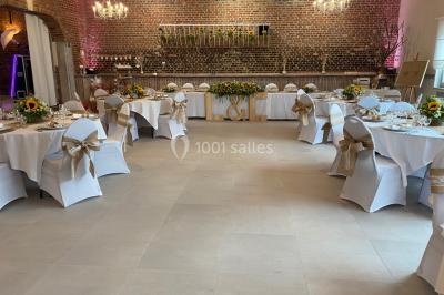 Salle de réception avec murs en briques, poutres apparentes, tables hautes et chaises pliantes sous des lampes suspendues.