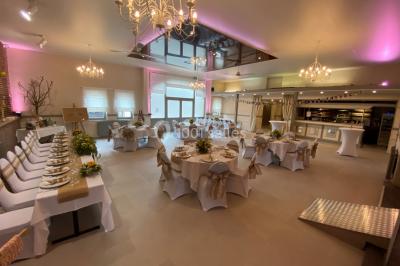 Salle de réception avec murs en briques, poutres apparentes, tables hautes et chaises pliantes sous des lampes suspendues.