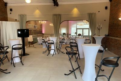 Salle de réception avec murs en briques, poutres apparentes, tables hautes et chaises pliantes sous des lampes suspendues.