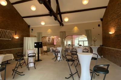 Salle de réception avec murs en briques, poutres apparentes, tables hautes et chaises pliantes sous des lampes suspendues.