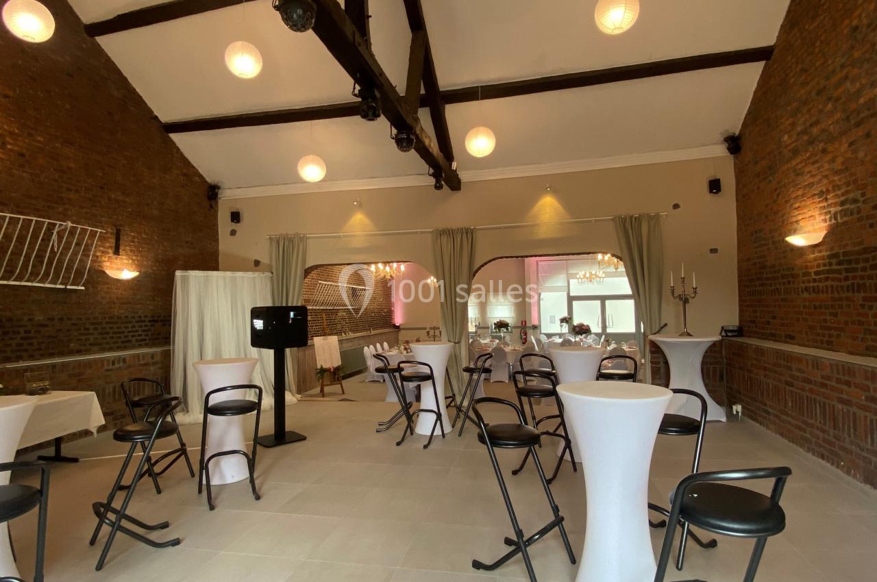 Salle de réception avec murs en briques, poutres apparentes, tables hautes et chaises pliantes sous des lampes suspendues.