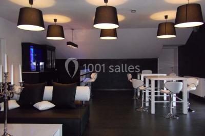 Salle de réception avec murs en briques, poutres apparentes, tables hautes et chaises pliantes sous des lampes suspendues.