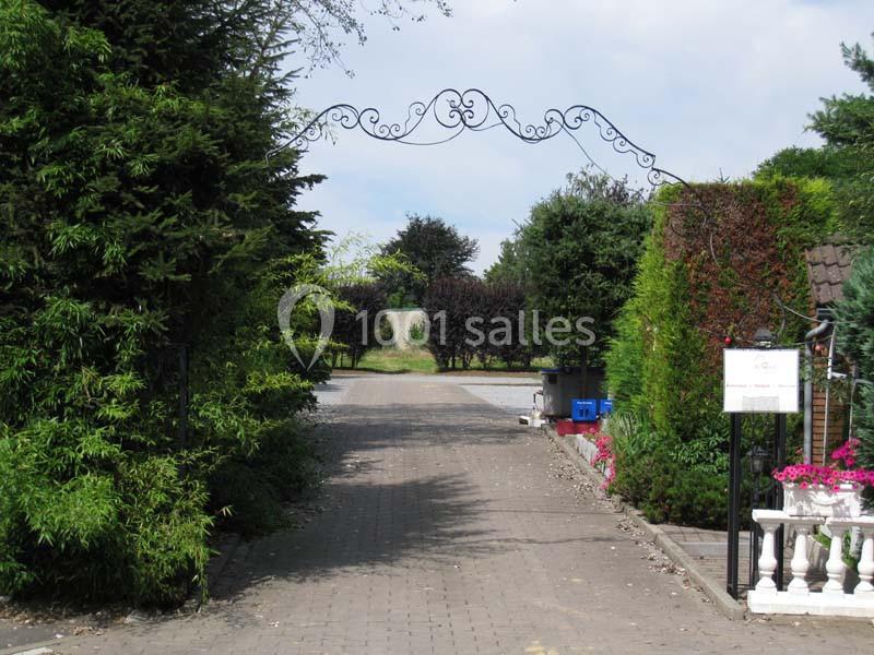 Allée pavée bordée de haies et de fleurs, avec une arche métallique décorative au-dessus de l'entrée.