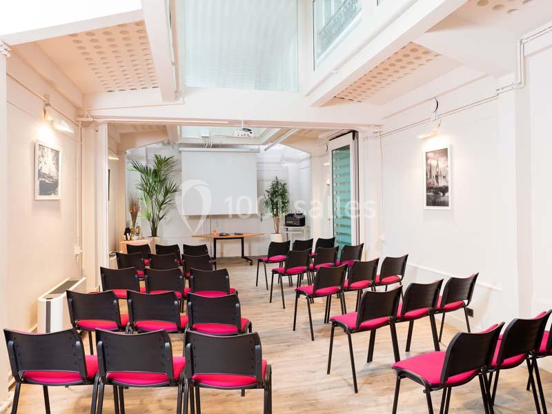 Salle lumineuse aménagée avec des chaises noires à assise rouge, un écran de projection et des plantes décoratives.